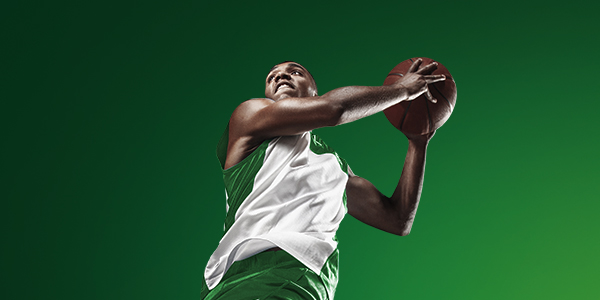 Basketballer in groene en witte outfit springt richting de basket met bal in de handen